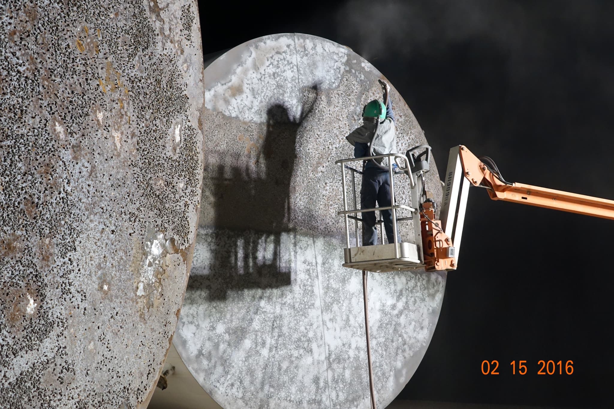 A worker in a boom lift basket blasting the curved wall of a large storage tank
